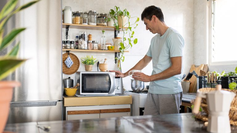 Eine Person steht in einer hellen K&uuml;che neben einer Mikrowelle und h&auml;lt eine wei&szlig;e Tasse in der Hand, w&auml;hrend sie auf das Ger&auml;t blickt.
