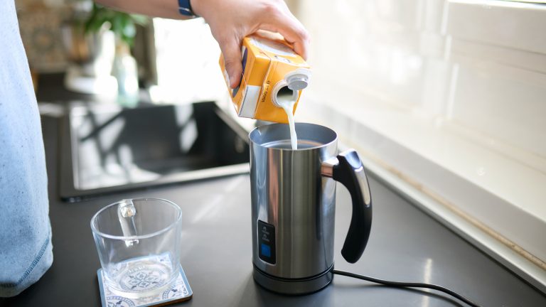 Eine Person f&uuml;llt Milch aus einem Milchkarton in einen elektrischen Milchaufsch&auml;umer aus Edelstahl auf einer dunklen K&uuml;chenarbeitsplatte.