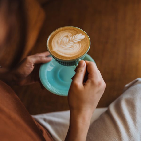 Eine Person h&auml;lt eine t&uuml;rkisfarbene Kaffeetasse mit kunstvoll gestaltetem Milchschaum in beiden H&auml;nden.