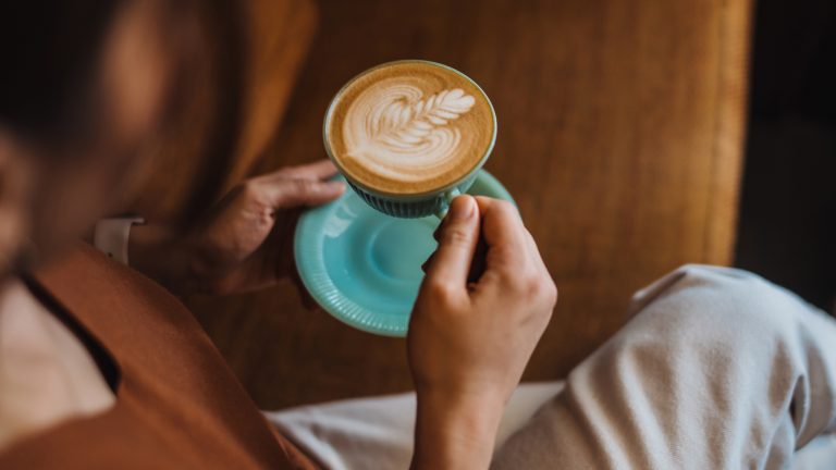 Eine Person h&auml;lt eine t&uuml;rkisfarbene Kaffeetasse mit kunstvoll gestaltetem Milchschaum in beiden H&auml;nden.