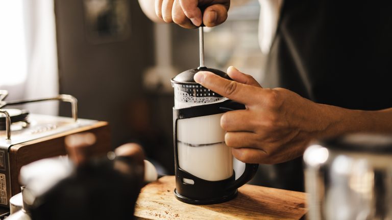 Eine Person pumpt mit einem French-Press-Milchaufsch&auml;umer Milch manuell auf, der Beh&auml;lter steht auf einem Holzbrett in einer rustikalen K&uuml;che.