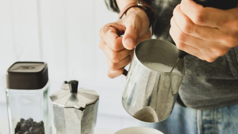 Eine Person sch&auml;umt Milch in einem Edelstahl-Milchk&auml;nnchen mit einem manuelle Milchaufsch&auml;umer, daneben eine Mokkakanne und ein Glas Kaffeebohnen.