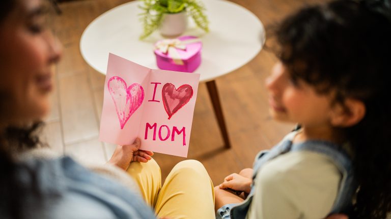 Ein Kind &uuml;bergibt seiner Mutter eine Karte auf der &bdquo;I love Mom&ldquo; steht. Im Hintergrund liegt eine kleine Geschenkbox.