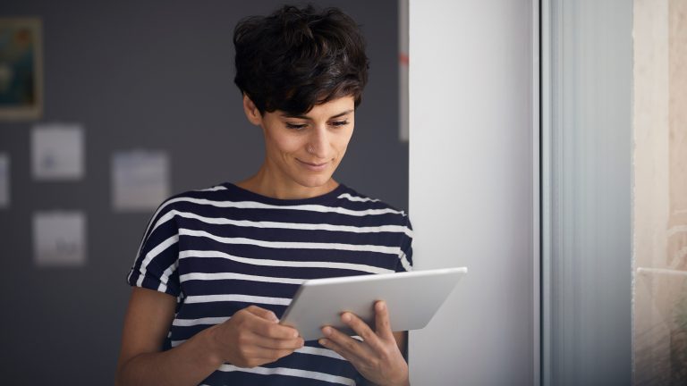 Eine Person lehnt an der Wand nahe eines Fensters und schaut auf ihr Tablet.
