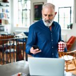 Eine Person steht mit einer Tasse in der Hand vor einem Laptop. In der Hand h&auml;lt sie ein iPhone.