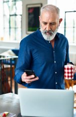 Eine Person steht mit einer Tasse in der Hand vor einem Laptop. In der Hand h&auml;lt sie ein iPhone.