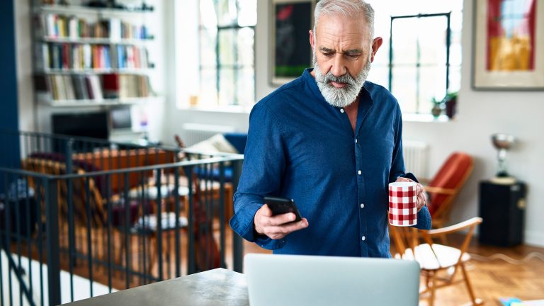 Eine Person steht mit einer Tasse in der Hand vor einem Laptop. In der Hand h&auml;lt sie ein iPhone.