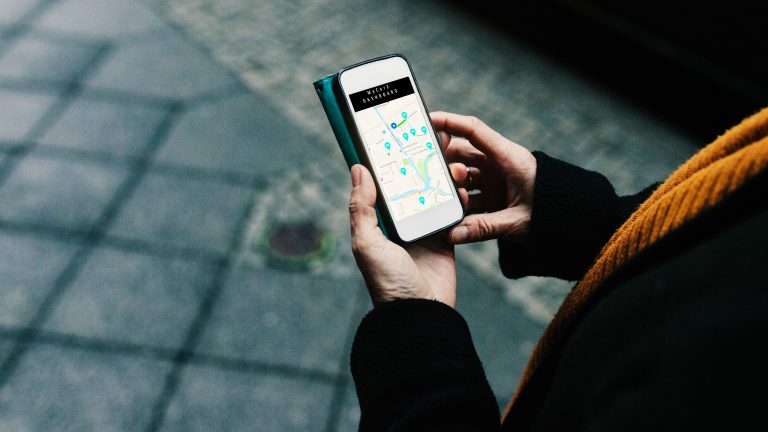 Eine Person auf einem Gehweg neben Autos schaut auf ihr Smartphone, auf dem nahe Carsharing-Autos angezeigt werden.