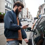 Eine Person steht neben einem Auto und scheint dieses &uuml;ber ihr Handy zu entsperren.