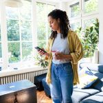 Eine Person steht in ihrem Wohnzimmer. In der einen Hand h&auml;lt sie eine Kaffeetasse, in der anderen ein Smartphone auf das sie gerade blickt.