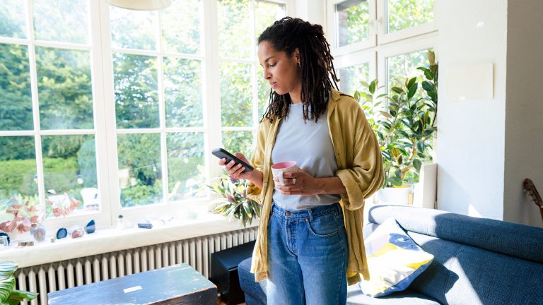 Eine Person steht in ihrem Wohnzimmer. In der einen Hand h&auml;lt sie eine Kaffeetasse, in der anderen ein Smartphone auf das sie gerade blickt.