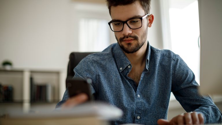 Eine Person sitzt an einem Schreibtisch und h&auml;lt dabei ihr Smartphone in den H&auml;nden.