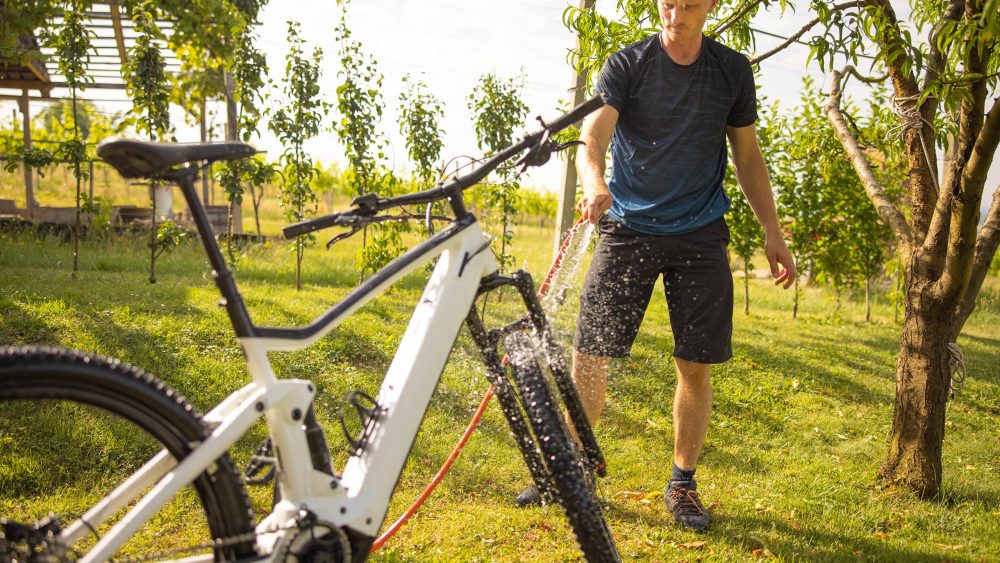 Eine Person steht auf einem Rasen und w&auml;scht ihr wei&szlig;es E-Mountainbike mit einem Gartenschlauch ab.