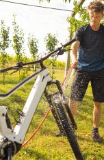 E‑Bike reinigen, So klappt es sicher und sauber Eine Person steht auf einem Rasen und wäscht ihr weißes E-Mountainbike mit einem Gartenschlauch ab.
