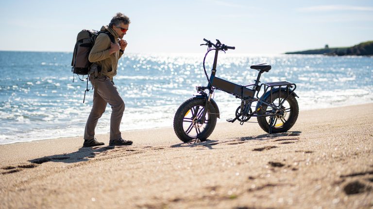 E‑Bike reinigen, So klappt es sicher und sauber Eine Person nähert sich einem klappbaren E-Bike an, das auf einem Sandstrand abgestellt ist.
