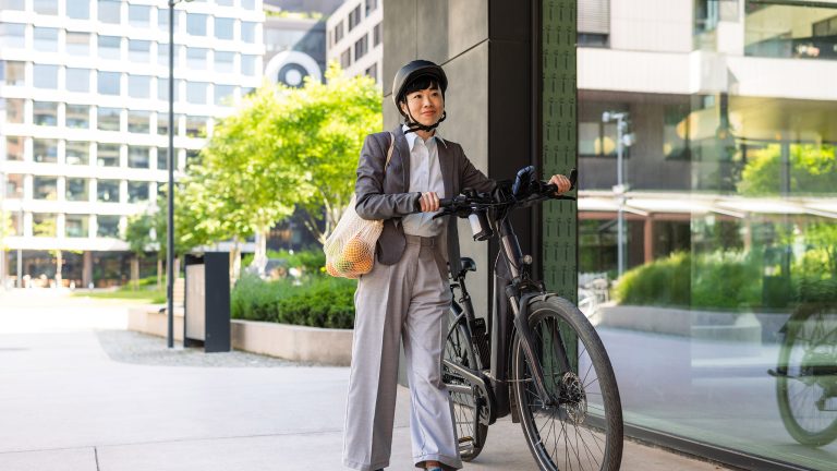 Eine Person, die einen Helm trägt und über der Schulter ihre Einkäufe, schiebt ein E-Bike durch die Stadt.