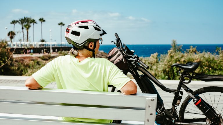 Rückansicht einer Person, die auf einer Bank sitzt und auf das Meer schaut. Vor der Bank steht ein E-Bike.