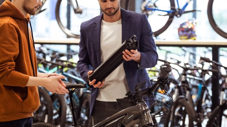 E-Bike-Akku defekt, Was du tun kannst, wenn die Batterie schwächelt Zwei Personen stehen in einem Fahrradgeschäft. Eine Person beäugt prüfend einen Akku, den sie in den Händen hält.