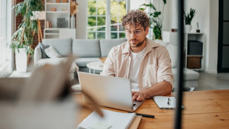 Eine Person sitzt an einem Tisch mit einem Laptop vor sich und scheint etwas zu tippen. Vor dem Laptop liegen einige Dokumente.