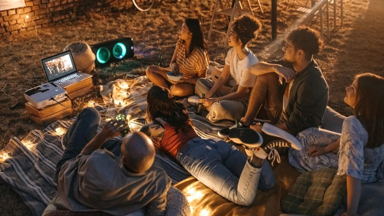 Eine Gruppe Leute sitzen auf einer Wiese auf einer Picknickdecke und schauen einen Film auf einem Laptop. Sie sind umgeben von einer warm-leuchtenden LED-Lichterkette.