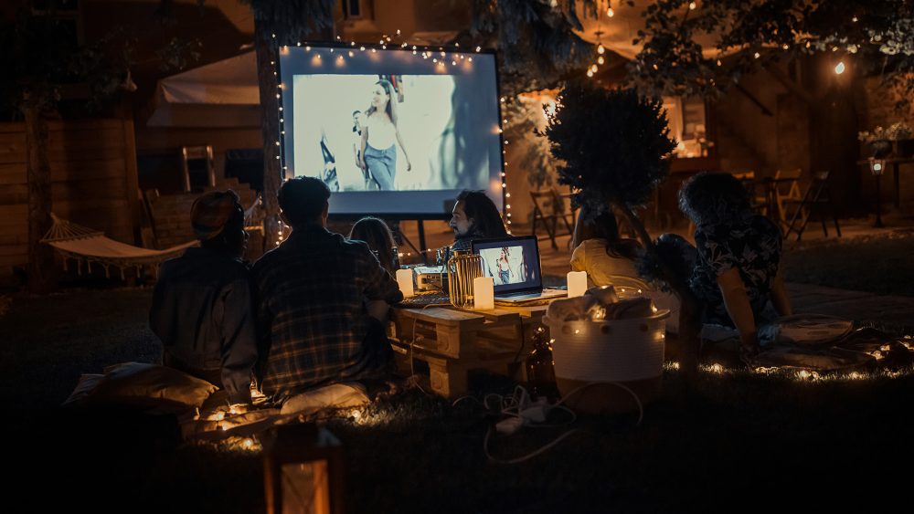 In einem Garten sitzen Leute, die einen Film auf einer Leinwand schauen. Es ist bereits dunkel, nur stimmungsvolles Licht umgibt sie.