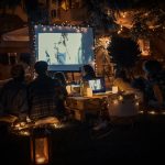 In einem Garten sitzen Leute, die einen Film auf einer Leinwand schauen. Es ist bereits dunkel, nur stimmungsvolles Licht umgibt sie.