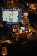 Outdoor-Kino, Mit diesen Gadgets wird dein Garten zum Lichtspielhaus In einem Garten sitzen Leute, die einen Film auf einer Leinwand schauen. Es ist bereits dunkel, nur stimmungsvolles Licht umgibt sie.
