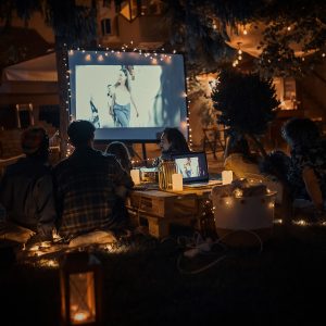 In einem Garten sitzen Leute, die einen Film auf einer Leinwand schauen. Es ist bereits dunkel, nur stimmungsvolles Licht umgibt sie.