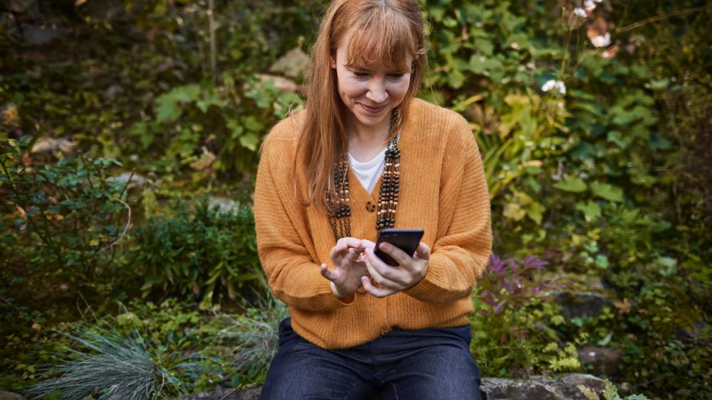 Eine Person sitzt auf einem Stein in einem Garten und schaut l&auml;chelnd auf ihr Smartphone.