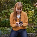 Eine Person sitzt auf einem Stein in einem Garten und schaut l&auml;chelnd auf ihr Smartphone.