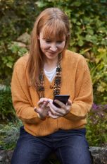 Eine Person sitzt auf einem Stein in einem Garten und schaut l&auml;chelnd auf ihr Smartphone.