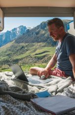 Überall vernetzt, Diese 5 mobilen Router bringen dich unterwegs ins Internet Eine Person sitzt im Bett ihres Campers und arbeitet am Laptop. Die Türen des Campers sind geöffnet und es ist ein Bergpanorama zu sehen.
