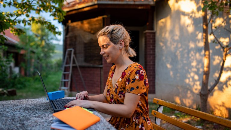 Eine Person sitzt auf einer Bank im Garten. Auf dem Tisch vor ihr steht ein Laptop.