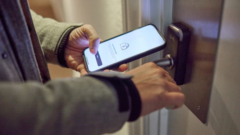 Detailaufnahme des Smartphones einer Person, die vor einer T&uuml;r mit einem smarten Schloss steht. Auf dem Screen des Phones ist eine App zum &Ouml;ffnen des Schlosses zu sehen.