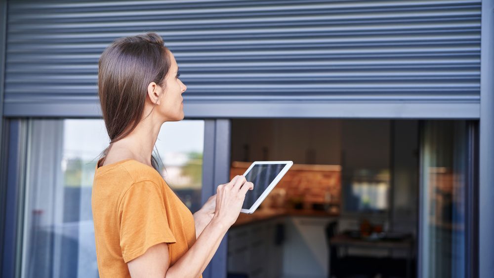 Eine Person h&auml;lt ein Tablet in der Hand und schaut dabei von au&szlig;en auf die Rolll&auml;den an einem Fenster.