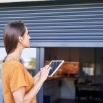 Eine Person h&auml;lt ein Tablet in der Hand und schaut dabei von au&szlig;en auf die Rolll&auml;den an einem Fenster.