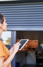 Smarte Rollladensteuerung, Was es ist und was du wissen solltest Eine Person hält ein Tablet in der Hand und schaut dabei von außen auf die Rollläden an einem Fenster.
