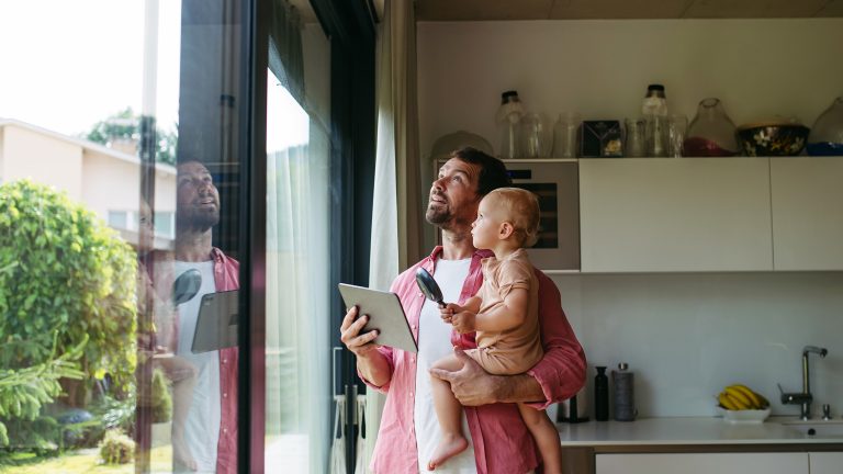Eine Person steht mit einem kleinen Kind auf dem Arm und einem Tablet in der Hand an einem gro&szlig;en Fenster.