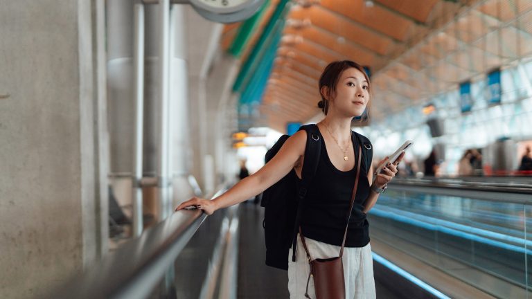 Eine Person steht auf einem Laufband im Flughafen. Mit der einen Hand h&auml;lt sie sich fest, in der anderen h&auml;lt sie ein Smartphone.