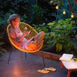Eine Person sitzt in einem gelben Acapulco-Chair auf einer stimmungsvoll beleuchteten Terrasse. In den H&auml;nden h&auml;lt sie einen Drink und ihr Smartphone.