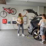 Eine Person schlie&szlig;t den Kofferraum von einem Auto. An dessen Ladeanschluss steht eine jugendliche Person, die gerade das Kabel der Wallbox am Auto anschlie&szlig;t.