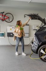Wallbox installieren, Modelle, Kosten und mehr Eine Person schließt den Kofferraum von einem Auto. An dessen Ladeanschluss steht eine jugendliche Person, die gerade das Kabel der Wallbox am Auto anschließt.
