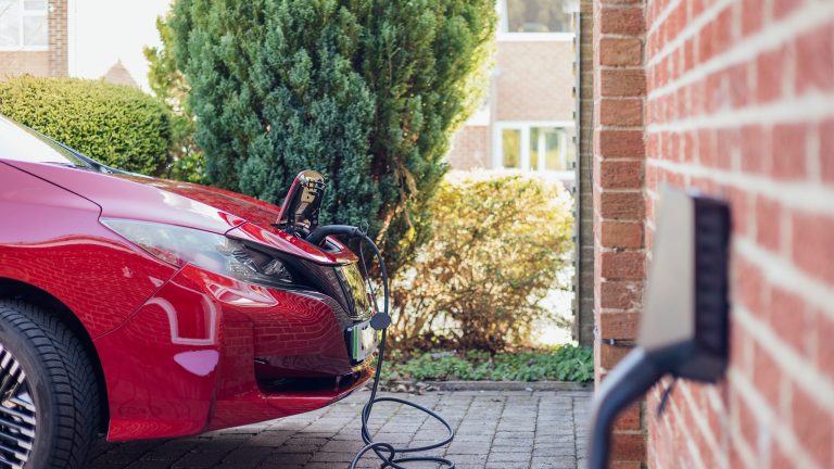 Ein rotes Elektroauto steht in einer Einfahrt und l&auml;dt an einer an einer Au&szlig;enwand angebrachten Wallbox auf.