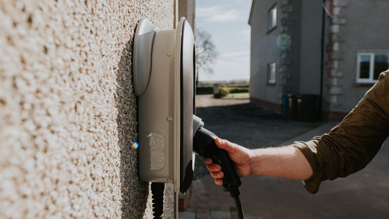 Eine Person hat die Hand am Anschlusskabel einer Wallbox, die an der Au&szlig;enwand eines Geb&auml;udes h&auml;ngt.