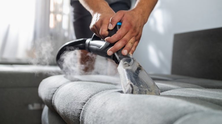 Dampfreiniger, Tipps und Tricks für die gründliche Reinigung Eine Person hält einen Dampfreiniger auf die Oberfläche eines Polstermöbels, während Dampf aus dem Gerät austritt.