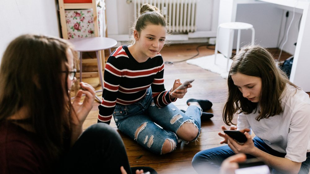 Drei junge Personen sitzen auf dem Boden in einem Zimmer. Alle drei halten ein Smartphone in der Hand.