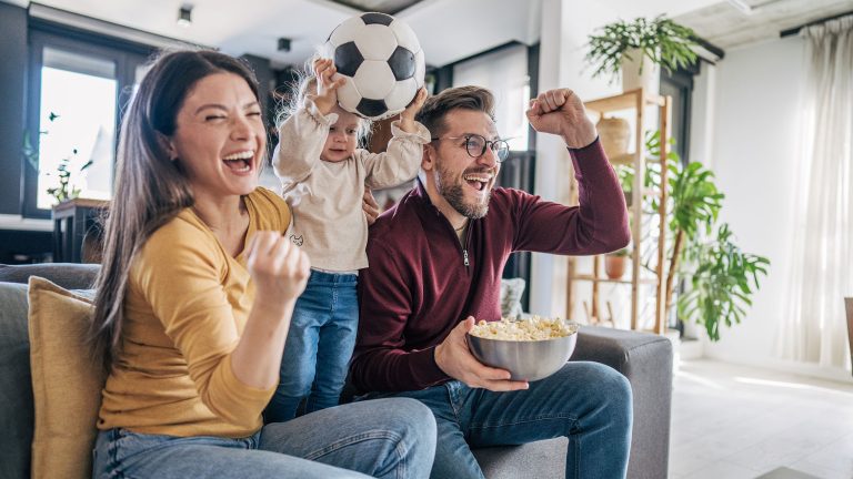 Zwei Personen und ein Kind sitzen auf einem Sofa und jubeln. Eine Person h&auml;lt Popcorn in der Hand, das Kind einen Fu&szlig;ball in die Luft.