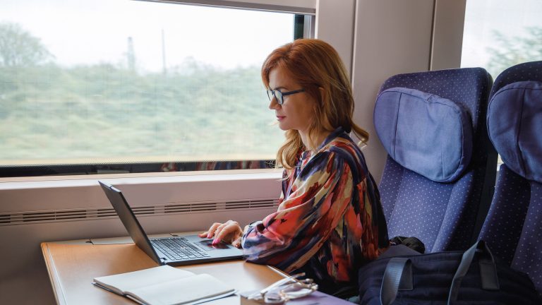 Eine Person sitzt in einem Zug und bedient einen Laptop vor sich. Daneben liegen B&uuml;cher.