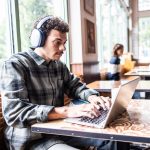 Eine Person sitzt mit Apple AirPods Max auf dem Kopf an einem MacBook Pro in einem Caf&eacute;.