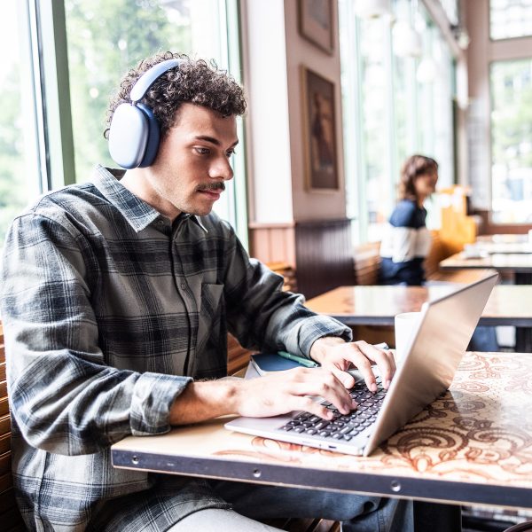 Eine Person sitzt mit Apple AirPods Max auf dem Kopf an einem MacBook Pro in einem Caf&eacute;.